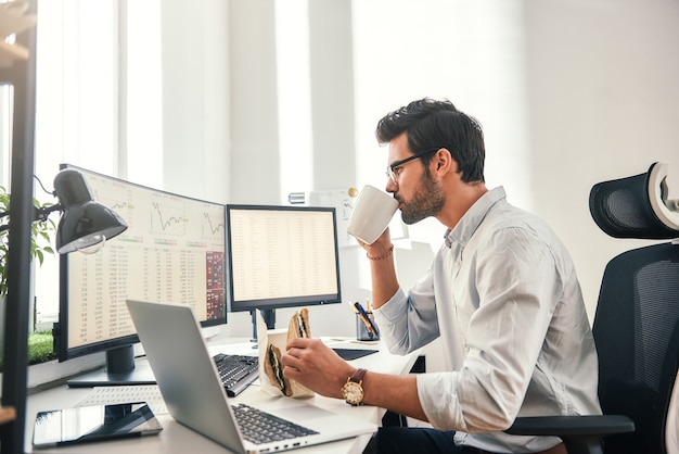 Coffee break young bearded businessman or trader is drinking a coffee and eating a sandwich