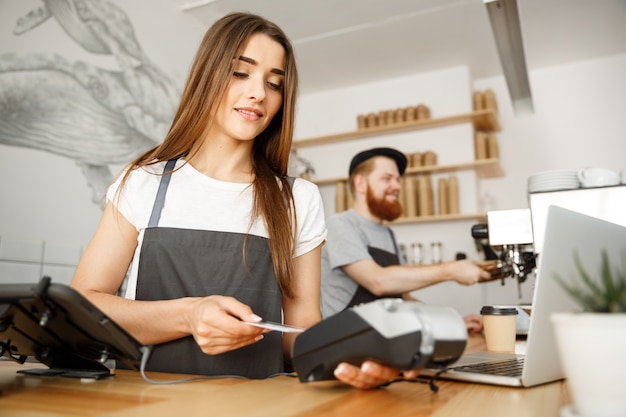 Coffee Business Concept - Beautiful female barista giving payment service for customer with credit card and smiling while working at the bar counter in modern coffee shop.