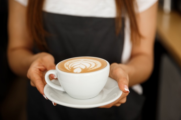 Coffee Business Concept - Cropped Close up of female serving coffee with latte art while standing in coffee shop. Focus on female hands placing a cup of coffee.