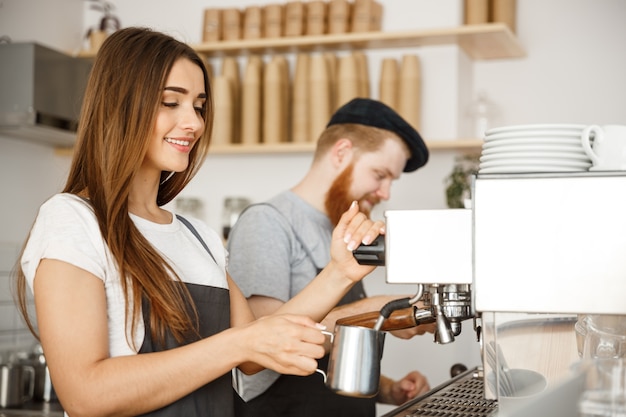 Coffee Business Concept - portrait of lady barista in apron preparing and steaming milk for coffee order with her partner while standing at cafe.