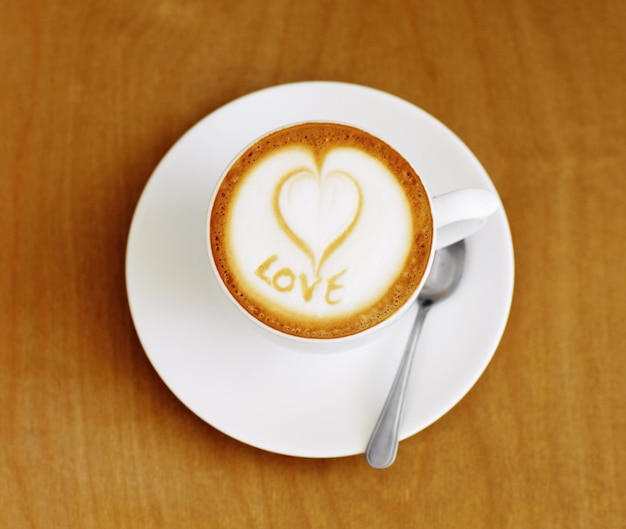 Photo coffee cup cappuccino and heart in foam on a table with no people in a restaurant and diner cafe drink love writing and foamy beverage art with milk in a cafe with a closeup of mug and spoon