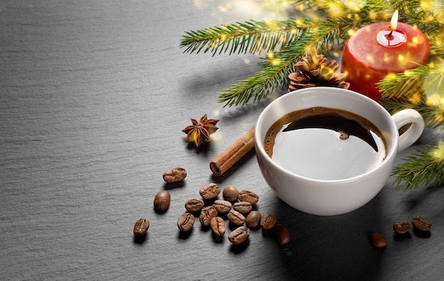 Coffee cup and coffee beans with christmas decoration fir branch and burning red candle on dark stone textured table.