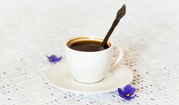 Coffee cup and saucer with purple African violet flowers on a white table surface.