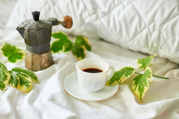 Coffee cup on white bed sheets with geyser coffee maker and green leaves