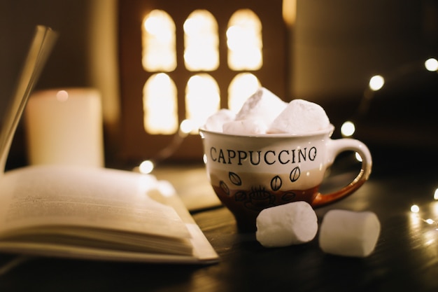 Coffee cup with marshmallows and a book on the table. Still life on dark background, flat lay