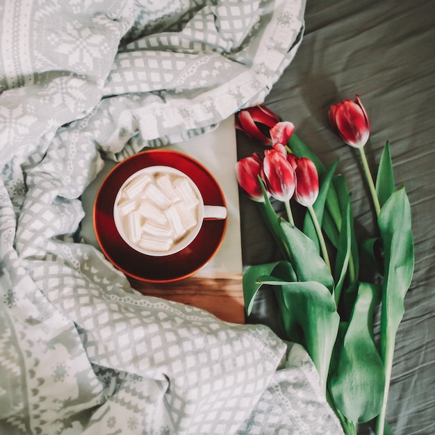 Coffee cup with marshmallows and red tulips in bed. Concept of holiday, birthday, Valentines Day, March 8