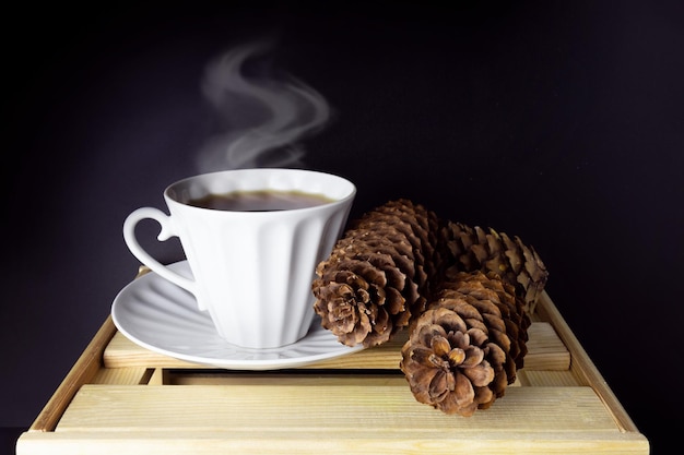 Coffee cup with steam and three fir cones on wooden table on dark background