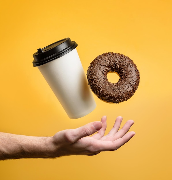 A coffee glass and a chocolate donut levitate above an outstretched palm on a yellow background