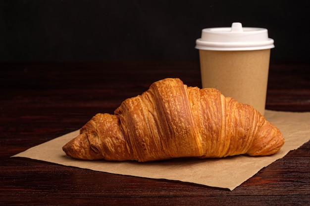 Coffee in paper cup and croissant on wooden table