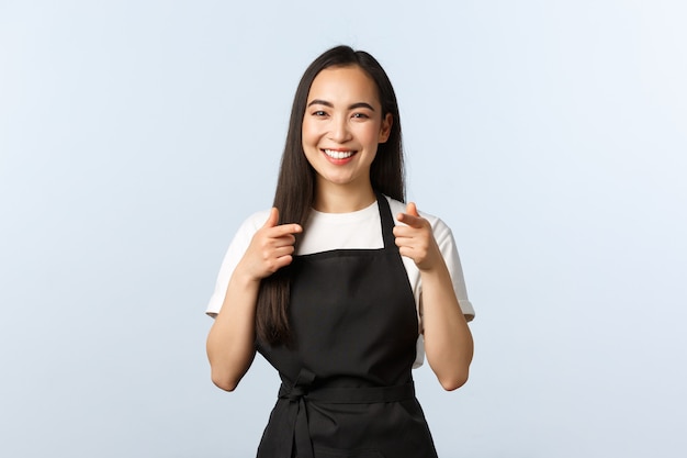 Coffee shop, small business and startup concept. Cheerful smiling female barista, asian cafe staff in black apron pointing at camera, inviting consumers visit and try new desserts, white background