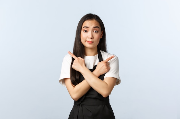 Coffee shop, small business and startup concept. Cute cafe employee in black apron asking advice as pointing sideways. Pretty asian waitress or barista showing left and right choices being indecisive.