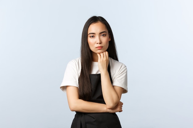 Coffee shop, small business and startup concept. Serious-looking thoughtful asian girl cafe owner thinking, hold fist under chin and stare camera as listening, standing white background