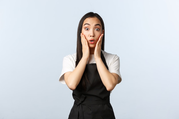 Coffee shop, small business and startup concept. Surprised asian girl barista in black apron, gasping in amazement, touch cheeks and stare camera astonished. Female cafe owner react to news