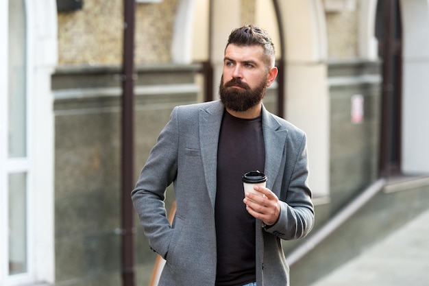 Coffee time. Businessman in hipster style holding takeaway coffee. Hipster with disposable paper cup walking in city. Bearded man enjoying morning coffee. Drinking his cup first thing in the morning.