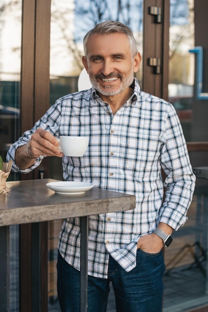 Coffee time. Cheerful middle aged man standing drinking coffee outdoors smiling confidently at camera during daytime