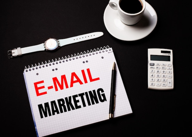 Coffee in a white cup, a wrist watch and a calculator on a black table. Nearby is a pen and a notebook with the text E-MAIL MARKETING