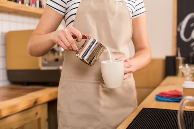 Coffee with milk. Close up of milk being poured in the cup by a pleasant delighted skillful woman while working in the coffee shop