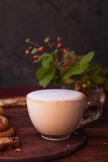 Coffee with milk foam in a glass mug on a wooden table next to a bouquet of wildflowers and cookies