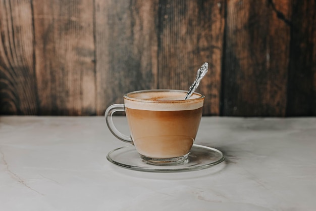 Coffee with milk in a transparent cup with a spoon on a plate on a dark wooden background