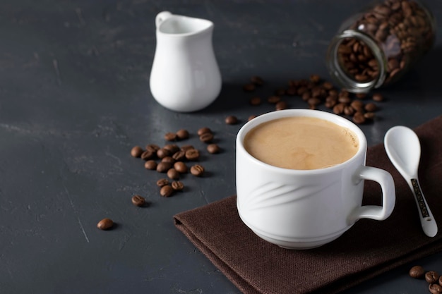 Coffee with milk in a white cup as well as scattered coffee beans on a gray table