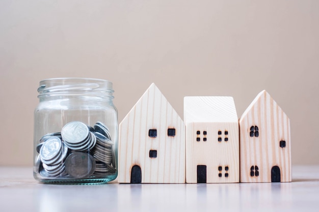 Coin in glass jar and wooden house model on table background.