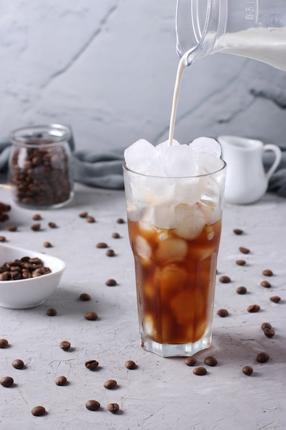 Cold coffee in a tall glass with cream being poured into it showing the texture of the drink Light gray background Vertical format Closeup