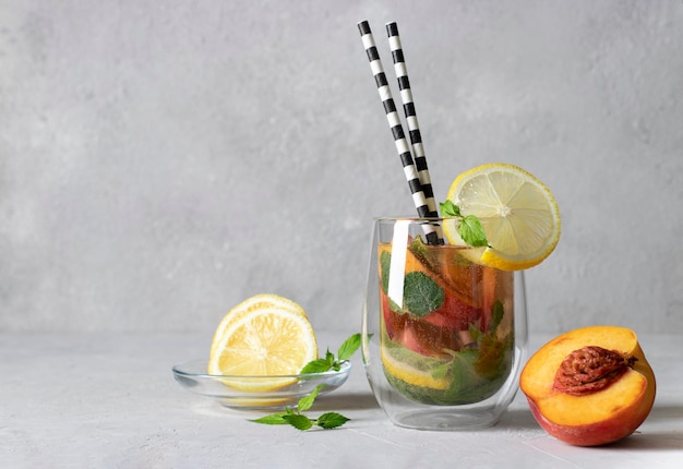 Cold mojito cocktail with peaches mint lemon slices and a drinking straw on light gray background