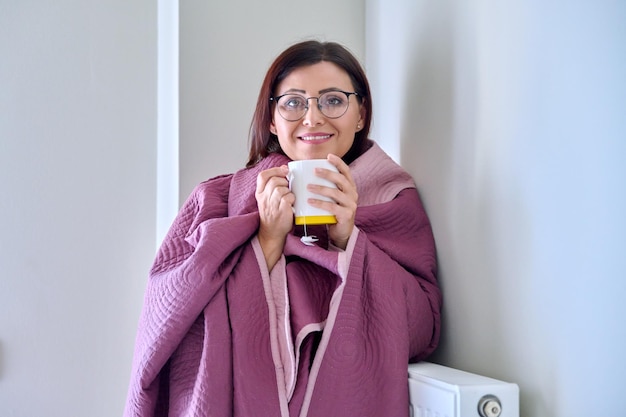 Cold season woman with plaid mug of hot tea warming near wall heating radiator