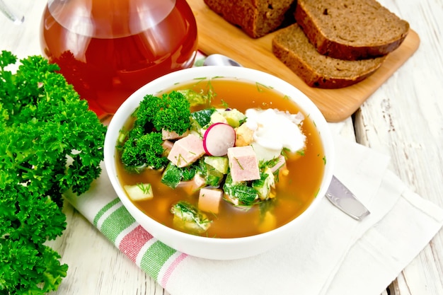 Cold soup okroshka from sausage, potatoes, eggs, radish, cucumber, greens and kvass in a white bowl on napkin, bread and jug with drink on the background of light wooden board