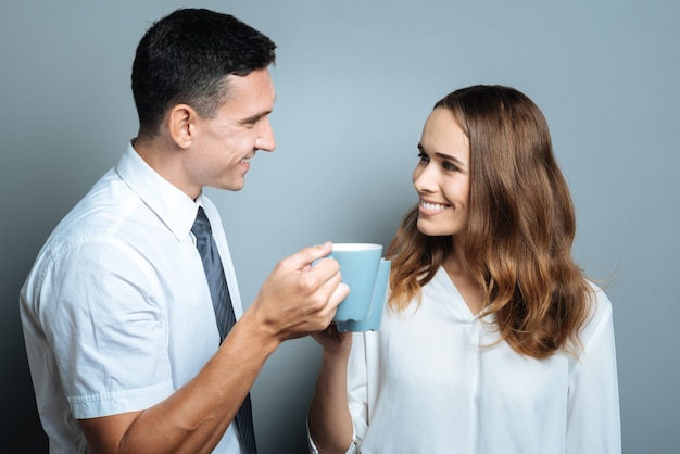 Colleagues at work. Joyful delighted positive people smiling and looking at each other while cheering with tea