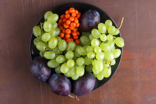 Collection of raw and organic autumn fruits from the garden closeup Ripe juicy grapes plum and rowan on a black plate on a dark background