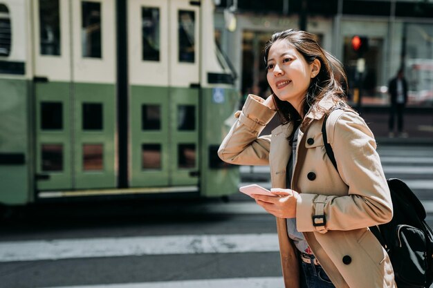 college girl study abroad sightseeing on weekend holidays. female teenager  holding cellphone searching plan route online map standing on road zebra crossing. green bus light train drive through.