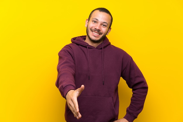 Colombian man with sweatshirt over yellow wall shaking hands for closing a good deal