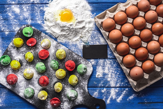 Colored dumplings on a bright blue wooden background, on a black board, flour is scattered next to a package of raw eggs, one egg is broken into flour