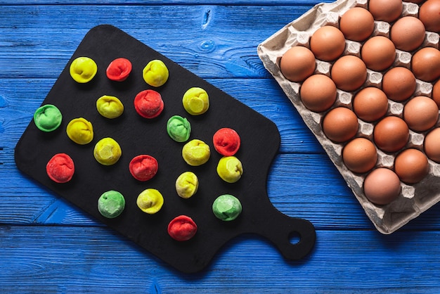 Colored dumplings on a bright blue wooden background, on a black board, next to a package of raw eggs