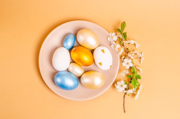 Colored eggs for Easter on a plate with branches of blossoming cherry on a beige background. Seasonality concept, spring, postcard, holiday. Flat lay, place for text. View from above.