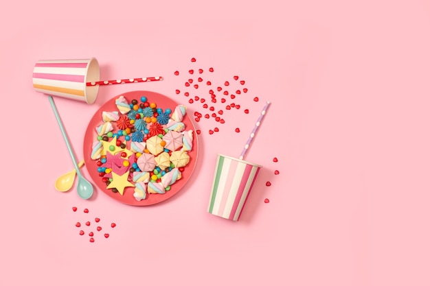 Colored meringues and candies in a pink plate and disposable cups on a pink background