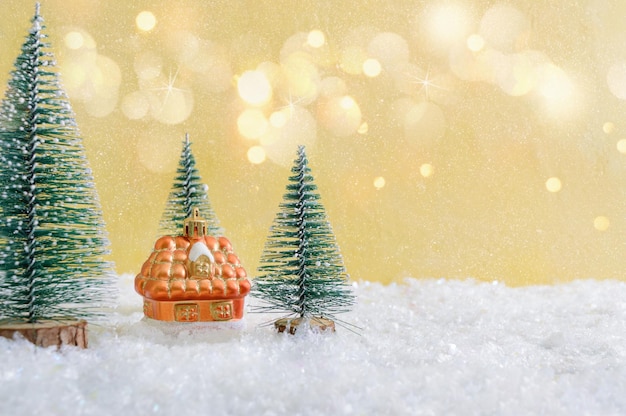 Colorful christmas toy house in the snow among the christmas trees. Background with festive lights. Cozy home buying concept. Beautiful Christmas card, copy space.