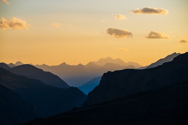 Colorful dawn landscape with beautiful blue mountains silhouettes and golden gradient sky with clouds. Vivid mountain scenery with picturesque multicolor sunset. Scenic sunrise view to mountain range.