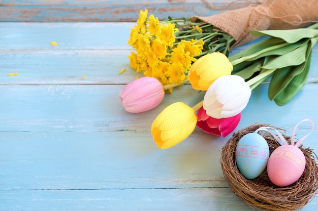 Colorful Easter eggs in nest with flowers on blue wooden background.  