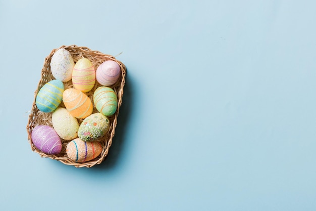 Colorful Easter eggs in wicker basket against colored background closeup top view with copy space