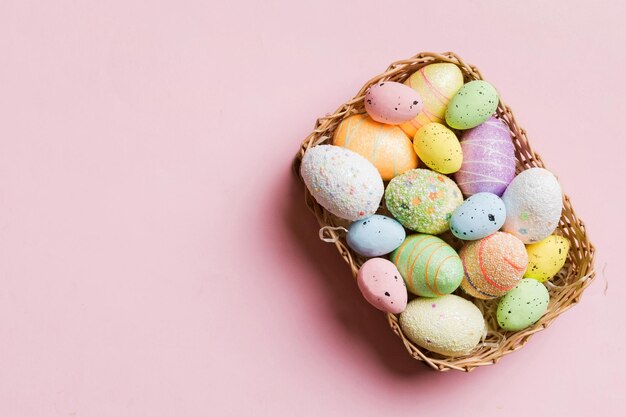 Colorful Easter eggs in wicker basket against colored background closeup top view with copy space