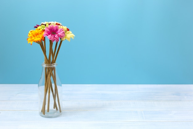 Colorful flowers in clear bottle on light blue table in front of blue wall. 