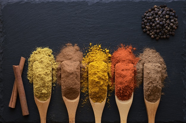 Colorful herbs and spices in wooden spoons on black stone table,  top view.