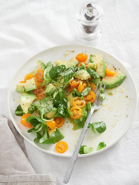 Colorful mixed salad of orange cherry tomatoes avocado and lamb's lettuce Plate with fork on table cloth next to pepper grinder