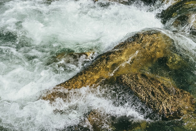 Colorful nature background with big boulder in turbulent flow of mountain river in sunny day. Water surface closeup with big stone in sunshine. Beautiful rapids in fast river. Mountain creek close up.