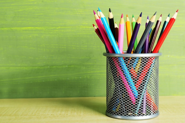 Colorful pencils in metal holder on wooden table and green wooden background