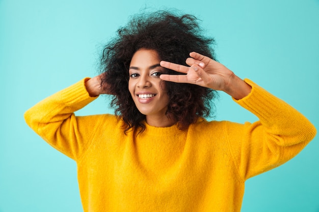 Colorful portrait of amazing woman with shaggy hair smiling and showing victory symbol with fingers, isolated over blue wall