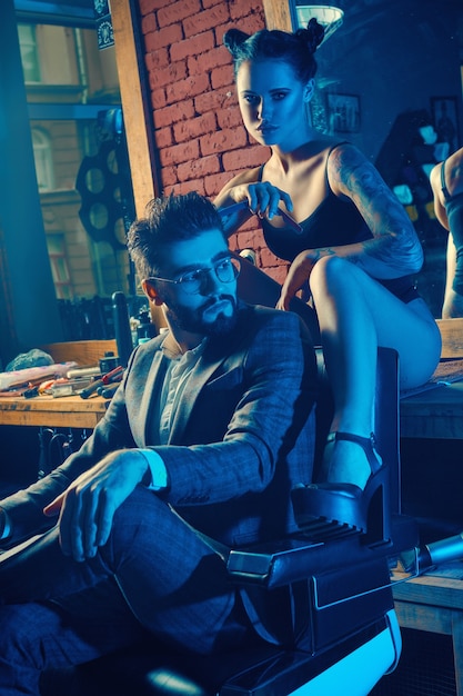 Colorful portrait of beautiful couple: brutal man in elegant suit and sexy girl with a tattoo wearing lingerie in barbershop