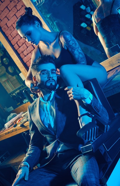 Colorful portrait of beautiful couple: brutal man in elegant suit and sexy girl with a tattoo wearing lingerie in barbershop